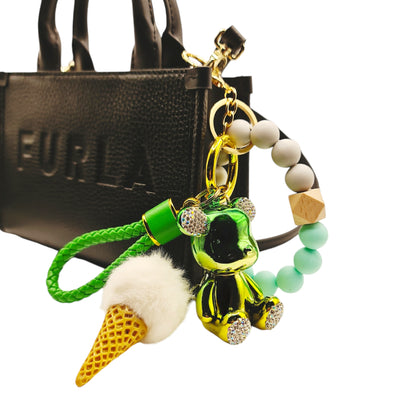 Cute and colorful ice cream-themed keychain featuring a pink pom-pom, a gold metallic teddy bear charm with diamond ears, and a beaded leather like strap. Featured on a Furla designer women's bag.